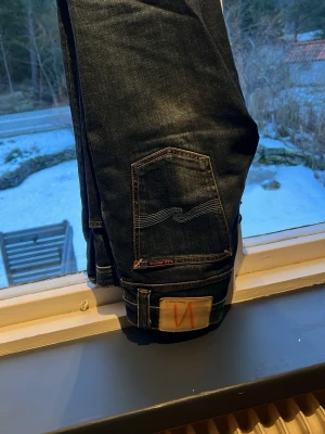 Mörkblå jeans från Nudie Jeans - Säljer ett par mörkblå jeans från Nudie Jeans med klassisk femficksdesign och kontrastsömmar. Jeansen har en snygg rak passform och är tillverkade i slitstarkt bomullsdenim.