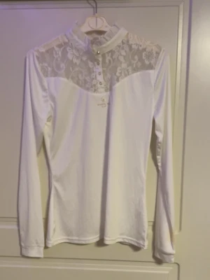 Vit långärmad spetsblus - Snygg vit blus med lång ärm och hög krage. Överdelen och ryggen har transparent spets med blommönster, vilket ger en romantisk vibe. Knäppning framtill med små knappar. Perfekt till jeans eller kjol för en trendig look.