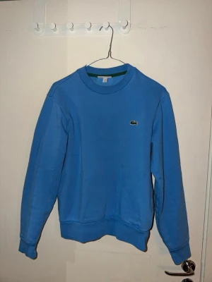 Blå sweatshirt från Lacoste (XS) - Säljer en klassisk blå sweatshirt från Lacoste med rund halsringning och den ikoniska krokodilloggan broderad på bröstet. Tröjan har ribbade muddar vid ärmslut och nederkant. Perfekt för en avslappnad och stilren look.