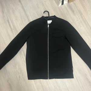 Svart jacka från Lindbergh - Snygg svart jacka från Lindbergh med dragkedja framtill och en bröstficka. Jackan har långa ärmar med knappar vid ärmsluten. Perfekt för en stilren look.