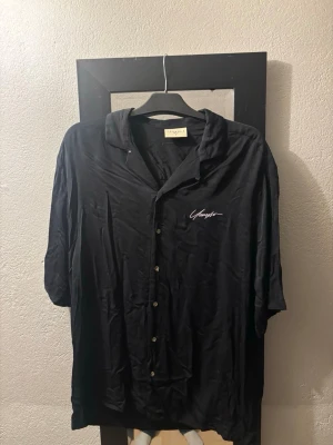 Svart skjorta från YoungLA - Svart skjorta från YoungLA med avslappnad passform och klassisk krage. Skjortan har knappar framtill och ett diskret broderat motiv på bröstet. Perfekt för en stilren och enkel look.