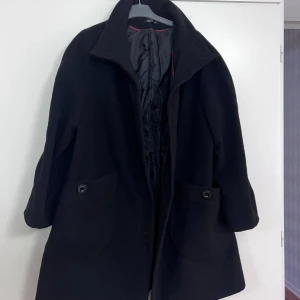 Svart kappa från XINT, st 48-50 - Stilren svart kappa från XINT i storlek XL. Klassisk design med stora fickor framtill, knäppning med knappar och hög krage. Fodrad insida med diskret röd kant. Perfekt för dig som vill ha en tidlös och snygg look under kyliga dagar.