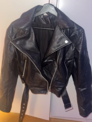 Svart biker skinnjacka med bälte - Snygg svart skinnjacka i klassisk bikerstil med bred krage, dragkedja och bälte i midjan. Jackan har långa ärmar och silverfärgade detaljer som ger en cool vibe. Perfekt för dig som vill ha en edgy look.