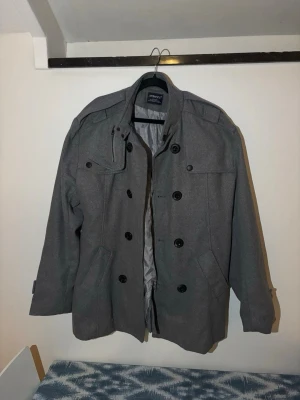 Grå dubbelknäppt rock från Jack & Jones - Stilren grå rock från Jack & Jones med dubbelknäppning och axelklaffar. Rocken har två stora fickor framtill, dekorativa knappar och klassisk krage. Perfekt för kyligare dagar och ger en snygg, tidlös look.