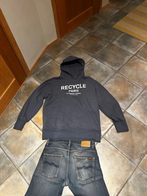 Mörkgrå hoodie Recycle Paris - Snygg mörkgrå hoodie från Superdry Studios med trycket 'RECYCLE PARIS' i vitt på bröstet. Klassisk huva och magficka framtill. Tillverkad i mjukt bomullsmaterial, perfekt för en avslappnad streetwear-look. Oanvänd eftersom att den var för stor