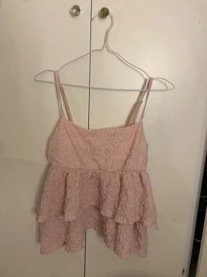 Rosa volangtop med tunna axelband - Söt ljusrosa linnetopp med volanger och tunna axelband. Toppen har en skrynklig textur och är luftig med dubbla lager tyg, perfekt för varma dagar. Ger en romantisk och trendig look.