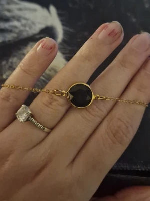 Guldfärgat armband med svart sten - Snyggt armband med tunn guldfärgad kedja och en rund, svart slipad sten i mitten. Stenen är infattad i en guldram som ger en elegant kontrast. Perfekt accessoar för att lyfta din outfit med en stilren detalj.
