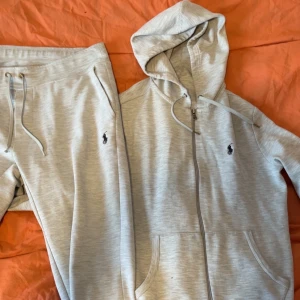 Grått loungewear-set från Polo Ralph Lauren - Mysigt loungewear-set i ljusgrått från Polo Ralph Lauren. Setet består av en zip-hoodie med dragsko och fickor samt matchande byxor med resårmidja och snörning. Båda delarna har den klassiska Polo-loggan broderad på bröstet och benet. 
