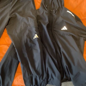 Svart Adidas träningsset/jacka  med huva - Säljer ett svart träningsset från Adidas med jacka och byxor. Jackan har huva, dragkedja och vita Adidas-loggor på bröstet och benet. Setet är gjort i ett lätt och sportigt material, perfekt för träning eller chill. Snygg och enkel design med diskreta detaljer.