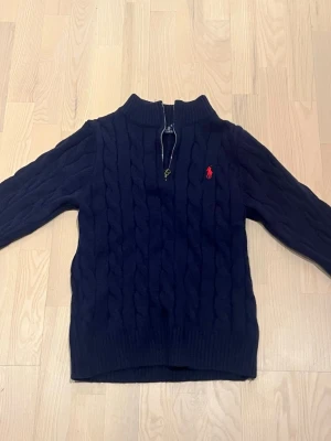 Mörkblå kabelstickad tröja Polo - Mörkblå kabelstickad tröja från Polo Ralph Lauren med halv dragkedja och ribbad krage. Tröjan har det klassiska röda Polo-logotypen broderad på bröstet och är gjord i mjukt material. Perfekt för en stilren och avslappnad look.