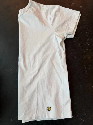 Vit t-shirt från Lyle & Scott - Klassisk vit t-shirt från Lyle & Scott med rund hals och korta ärmar. T-shirten har den ikoniska gula örnloggan broderad nere vid fållen. Tillverkad i mjuk bomull för en skön känsla och enkel stil.