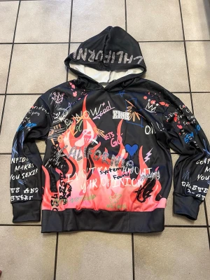 Svart hoodie med graffiti-tryck - Cool svart hoodie med färgglada graffiti-inspirerade tryck, flames och textdetaljer över hela plagget. Huva med texten 'CALIFORNIA' och insida i vitt. Tillverkad i ett mjukt, syntetiskt material som känns skönt mot huden. Perfekt för dig som gillar streetwear och unika plagg.