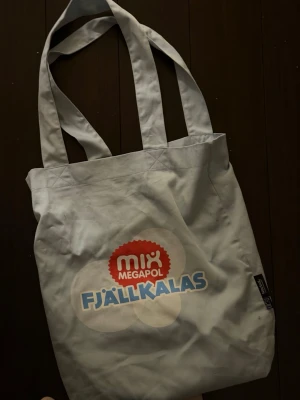 Ljusblå shoppingväska Fjällkalas - Ljusblå shoppingväska från Neutral med tryck 'Mix Megapol Fjällkalas' på framsidan. Tillverkad i 100% ekologisk och Fairtrade-certifierad bomull. Väskan har långa axelband och är perfekt för att bära med sig allt man behöver under dagen.