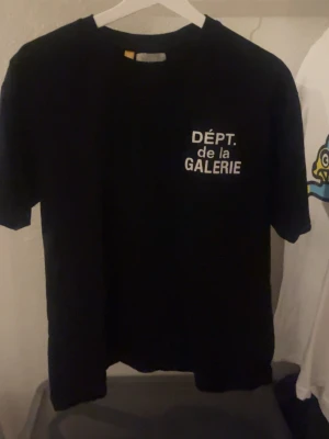 Svart t-shirt från Gallery Dept. - Säljer en svart t-shirt från Gallery Dept. med vit text 'DÉPT. de la GALERIE' tryckt fram och stort på ryggen. Klassisk passform och rund hals. Tillverkad i mjuk bomull, perfekt för en clean och stilren look. Tveka inte på att ställe en fråga eller två!😊😊🥰