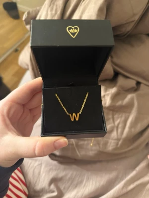 Guldhalsband med bokstaven W - Snyggt halsband i guld från ur&penn med ett hängsmycke i form av bokstaven W. Kedjan är tunn och elegant, och halsbandet levereras i en svart ask med hjärtdetalj. Perfekt för dig som vill ha en personlig touch på din stil.