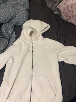 Vit hoodie med dragkedja Jack & Jones - Säljer en vit hoodie från Jack & Jones med dragkedja framtill och stora fickor. Hoodien har en klassisk huva med dragsko och är gjord i mjukt, fleecefodrat material som känns riktigt skönt mot huden. Perfekt för dig som gillar enkel och clean stil.