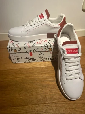 Dolce & Gabbana vita sneakers med röd detalj - Tjeneritz! Säljer ett par schysstare Dolce & Gabbana sneakers med röda detaljer på häl och plös. Skorna har inte använts oförsiktigt, och är i fantatiskt skick. Har ni fler frågor eller funderingar är det bara höra av er!      OBS andra bilden är lite förlängd då den är tagen i 0,5!!