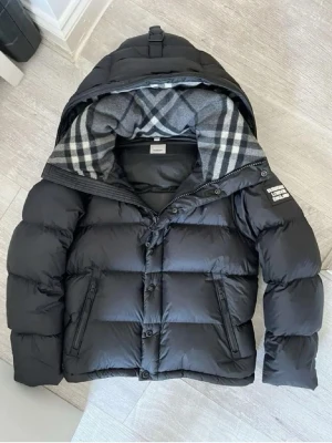 Burberry Jacka  - Säljer en svart pufferjacka från Burberry med ikoniskt rutigt foder i huvan. Jackan har dragkedja och knappar framtill, två sidofickor med dragkedja och justerbar huva. Tillverkad i polyester och dun, perfekt för kalla dagar.
