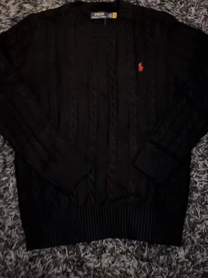 Svart  stickad tröja Polo Ralph Lauren - Kabelstickad svart  tröja från Polo Ralph Lauren med klassisk röd logga på bröstet. Rund halsringning och långa ärmar. Perfekt för dig som gillar tidlös och stilren design.När köparen köper varan skickas den samma dag eller dagen där på. 