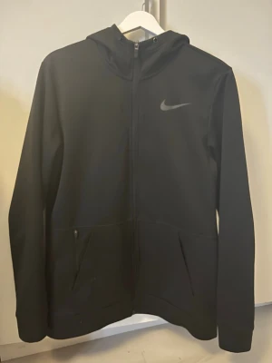 Svart Nike Therma-Fit hoodie - Säljer en svart Nike Therma-Fit hoodie med dragkedja framtill och två fickor. Hoodien har en stilren design med Nike-logga på bröstet och är tillverkad i ett mjukt, värmande material. 