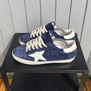 Blå Golden Goose Superstar sneakers - Golden Goose Superstar sneakers i blå mocka med vit stjärndetalj på sidan, vita snören och vit sula. Klassisk låg modell med rund tå och retrovibbar. Märkeslogga på hälen och tungan. Perfekta för dig som gillar streetstyle och unika detaljer.