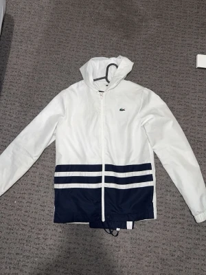 Vit och marinblå tracksuit Lacoste - Snygg vit vindjacka från Lacoste med marinblåa detaljer och två horisontella ränder nedtill. Jackan har huva, dragkedja framtill och justerbar nederkant med snörning. Perfekt för blåsiga dagar och sportig stil.