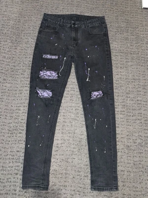 Svarta skinny jeans med lila detaljer - Säljer ett par svarta skinny jeans med slitningar och lila spetsdetaljer bakom hålen. Jeansen har även vita och lila färgstänk för en unik look. Perfekta för dig som vill sticka ut lite extra.