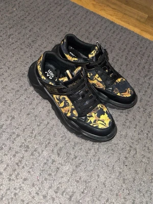 Versace Jeans Couture sneakers svart/guld - Chunky sneakers från Versace Jeans Couture i svart med guldigt barockmönster. Skorna har snörning, tjock sula och detaljer i syntetmaterial. Logga på sidan och plös för extra statement.