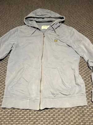Ljusblå hoodie från Lyle & Scott - Ljusgrå hoodie med dragkedja från Lyle & Scott. Klassisk gul logga på bröstet, två stora fickor framtill och justerbar huva med snören. Tillverkad i mjukt bomullsmaterial och har ribbade muddar vid ärmslut och nederkant.