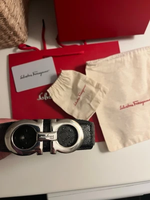 Svart Ferragamo bälte med silverdetaljer - Snyggt svart bälte från Salvatore Ferragamo i präglat läder med ikoniskt dubbel-Gancini-spänne i silverfärgad metall. Kommer med originalpåse och förpackning. Skick: använt fåtal gånger (9/10). Nypris: 4199kr. Det är pappas gamla bälte och hjälper han att bli av med det! Fråga gärna vid funderingar!