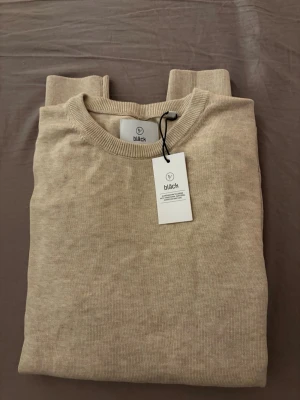 Beige stickad tröja från Bläck - Snygg beige stickad tröja från Bläck med rund halsringning och ribbade muddar vid ärmslut och nederkant. Tröjan har en stilren och enkel design som passar perfekt till lager-på-lager och är tillverkad i mjukt material.