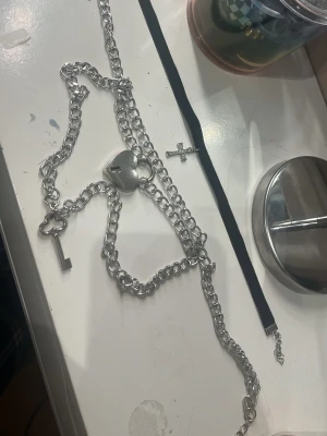 Två halsband med lås och kors - Säljer två halsband: ett chunky silverfärgat kedjehalsband med hängande hjärtlås och nyckel. Sen ett svart sammetsband med ett litet silverfärgat kors. Perfekta accessoarer för en edgy och alternativ stil.