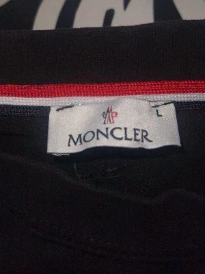 Svart Moncler sweatshirt  - Svart sweatshirt från Moncler med stor broderad logga i rött och vitt på bröstet. Tröjan har rund halsringning med röd och vit randig detalj på insidan samt långa ärmar och ribbade muddar. Perfekt för en stilren och sportig look. Andvänts 1 gång 