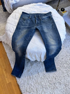 G-Star RAW 3301 Slim Jeans 28/32 - Säljer ett par blå G-Star RAW 3301 Slim jeans i storlek 28/32. Jeansen har klassisk femficksdesign, snygga slitningar och en skinnpatch bak i midjan. De är tillverkade i slitstarkt bomullsmaterial och har en smal passform som sitter snyggt längs benen. Lagad i grenen av skräddare annars bra skick 