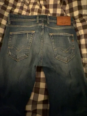 Replay blå jeans med eftertraktade slitningar - Säljer ett par blå Replay jeans med snygga slitningar och tvättade detaljer. Klassisk femficksmodell med raka ben och slim fit passform. Jeansen har läderpatch bak i midjan och coola sömmar på bakfickorna. Perfekt om du gillar jeans med lite slimare fit