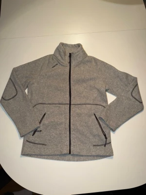 Grå fleecekofta från Tribord - Mysig grå fleecekofta från Tribord med hel dragkedja framtill och två praktiska fickor med dragkedja. Hög krage och markerade sömmar ger en sportig look. Perfekt för kyliga dagar och enkel att slänga på sig över en t-shirt. För liten för mig.