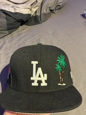 Svart LA New Era keps med palmer - Säljer en svart New Era keps med LA-logga framtill och broderade palmer i grönt och brunt på sidan. Kepsen har rak skärm och klassisk passform. MLB-logga broderad bak. Perfekt för dig som gillar streetwear och sportig stil.