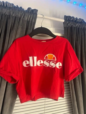 Röd croppad topp från Ellesse - Säljer en röd croppad topp från Ellesse med stort vitt logotryck framtill och en orange detalj. Toppen har korta ärmar och rund halsringning. Perfekt för dig som gillar sportig och avslappnad stil.