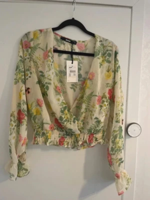 Blommig blus från Bik Bok - Söt blommig blus från Bik Bok. Blusen har lång ärm med resår vid ärmslut och volangdetalj i midjan. Perfekt för en fräsch och somrig look. Oanvänd med prislapp kvar. Går att knäppa framsidan (bild 2).