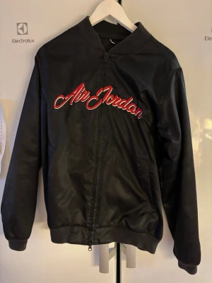 Svart Air Jordan bomberjacka - Snygg svart bomberjacka från Air Jordan med röd och vit broderad logga på bröstet. Jackan har dragkedja framtill, ribbade muddar och klassisk bomberkrage. Perfekt för dig som vill ha en sportig och stilren look.
