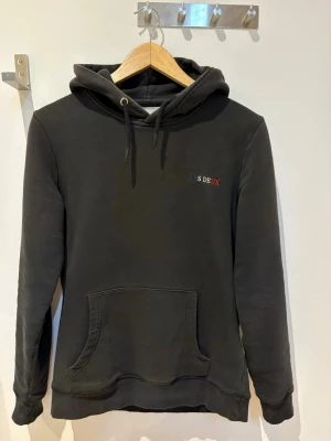 Svart hoodie från Les Deux - Svart hoodie från Les Deux med broderad logga på bröstet i vitt, blått och rött. Klassisk modell med huva, dragsnören och känguruficka framtill. Mjuk insida och ribbade muddar vid ärmslut och nederkant. Perfekt för en avslappnad stil.