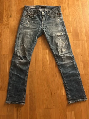 Replay slitna blå jeans herr - Snygga blå jeans från Replay med slitna detaljer och raka ben. Jeansen har klassisk femficksdesign och tydliga slitningar framtill för en cool, avslappnad look. Perfekta för dig som gillar streetstyle och vill ha ett par jeans med attityd. Jeansen är lite skitiga samt tecken på användning vid höger bakficka (se bild) Storlek 29.30