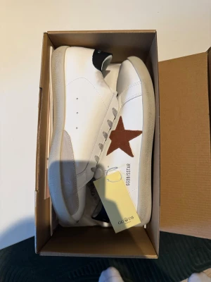 Vita Golden Goose sneakers med stjärna - Golden Goose sneakers i vitt skinn med grå snörning och en brun stjärna på sidan. Skorna har rund tå, platt sula och svart detalj på hälen. Klassisk och stilren design med GGDB-logga på plösen.