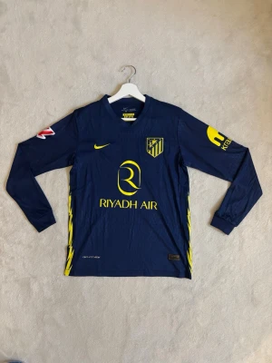 Atlético Madrid matchtröja - Mörkblå långärmad matchtröja från Atlético Madrid med gula detaljer och tryck. Nike-logga på bröstet, klubbmärke och sponsortryck 'Riyadh Air' framtill. 