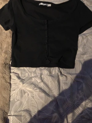 Svart croppad topp från 157 - Snygg svart croppad topp från 157 med korta ärmar och knappar framtill. Toppen har en rundad urringning och är gjord i mjuk bomull, perfekt att matcha med höga jeans eller kjol.