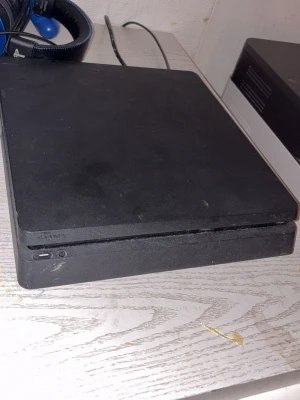 Sony PlayStation 4 Slim svart konsol + två kontroller och spel - Säljer en svart PlayStation 4 Slim från Sony. Konsolen har en tunn och stilren design med mattsvart finish och rundade hörn. Perfekt för gaming och streaming, med HDMI- och USB-portar på framsidan. Passar dig som vill ha en smidig spelupplevelse.