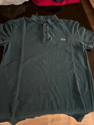 Mörkgrön pikétröja från Lacoste - Klassisk mörkgrön pikétröja från Lacoste med den ikoniska broderade krokodilloggan på bröstet. Tröjan har korta ärmar, krage och knappar framtill. Perfekt för en avslappnad men ändå stilren look.