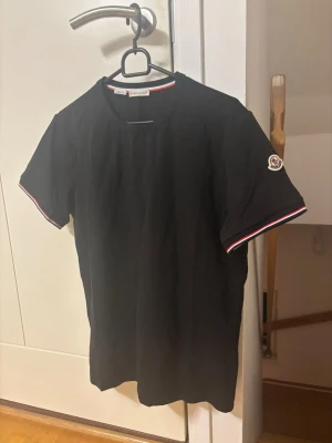 Svart t-shirt från Moncler - Svart t-shirt från Moncler med korta ärmar och rund hals. Ärmsluten har rödvita ränder och Moncler-logga på vänster ärm. Perfekt till en clean och stilren look.