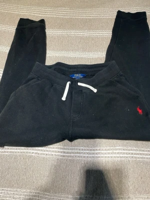 Svarta mjukisbyxor från Polo Ralph Lauren - Svarta mjukisbyxor från Polo Ralph Lauren med vit dragsko i midjan och klassisk röd logga broderad på benet. Byxorna har en relaxed passform och är tillverkade i mjuk bomull, perfekta för chill dagar eller när du vill vara extra bekväm. Storlek 158