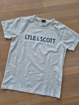 Vit Lyle & Scott t-shirt med tryck - Vit t-shirt från Lyle & Scott med stort rutigt logotyptryck över bröstet. Klassisk rund halsringning och rak passform. 95% bomull, 5% eldstad. Storlek L